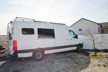A white Sprinter camper van with a roof rack and ladder, parked on gravel next to a house. Ideal for an RV rental adventure.
