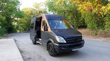 Matte black Mercedes Sprinter camper van, ideal for rv rental adventures, parked on a tree-lined road with its side door open.