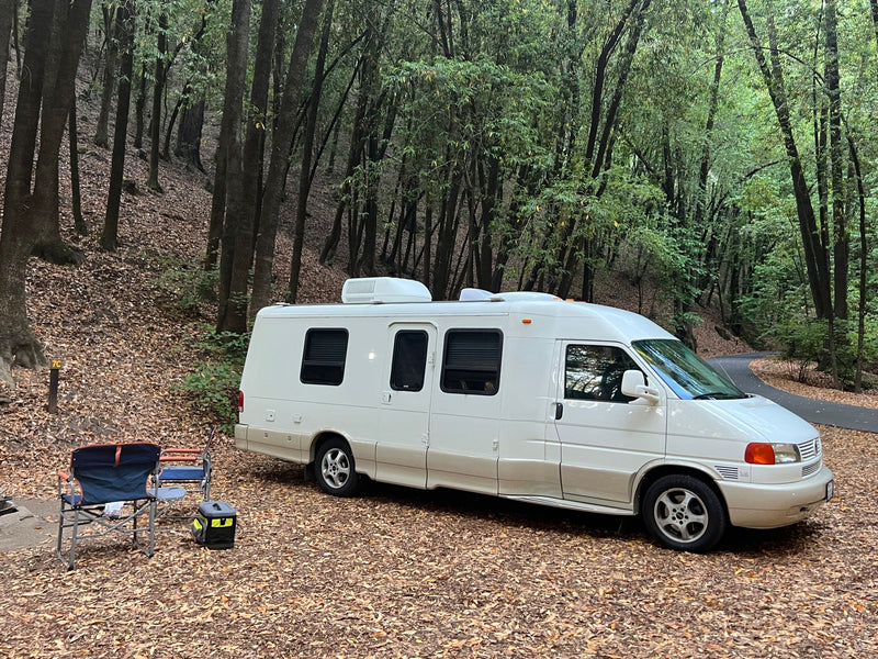  sunlit forest campsite.|RV rental parked in a tranquil forest campsite with chairs and cooler ready for a relaxing outdoor getaway.|This white rv rental is nestled in a shady — photo 2