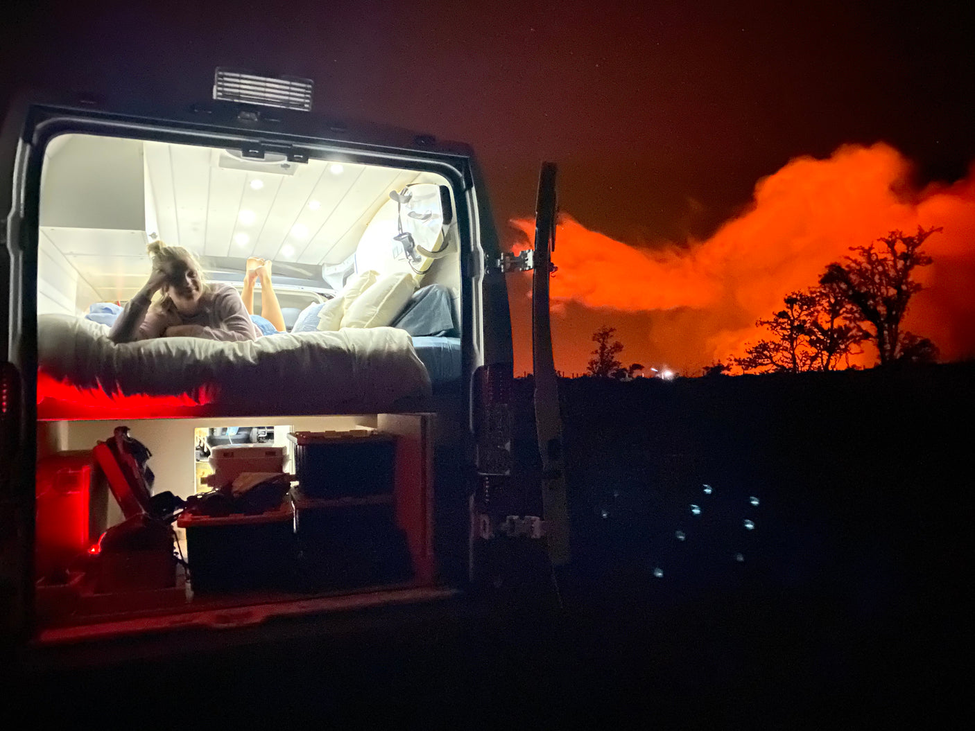 Inside an RV rental at night, a person rests in a cozy bed, contrasting with a dramatic orange glow (likely fire/smoke) illuminating the distant sky.