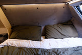 View inside an **rv rental**: a compact sleeping area with two olive green pillows, a white duvet, and overhead LED lighting.