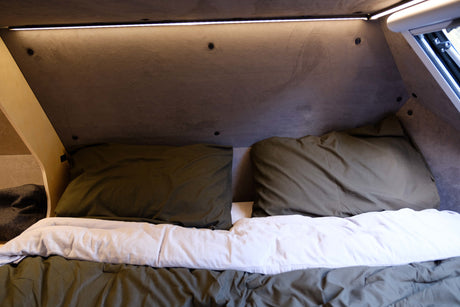 View inside an **rv rental**: a compact sleeping area with two olive green pillows, a white duvet, and overhead LED lighting.