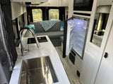 Modern RV rental interior: well-equipped kitchen with sink & cooktop, open fridge, and inviting living area with a comfy sofa.