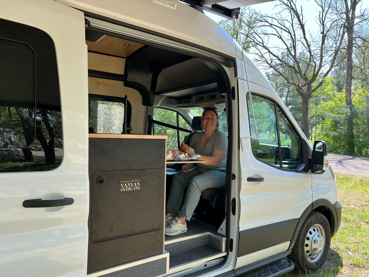 A smiling woman enjoys her burger inside a white Vanvan Overland RV rental, ready for travel.
