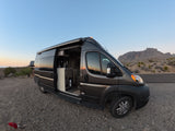 An RV rental campervan parked on gravel, showcasing its open side door, interior kitchenette, and a mountain desert sunset backdrop.