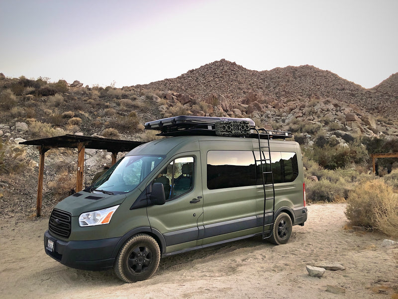 Green Ford Transit RV rental camper van parked in a dry, rocky desert campsite with a simple wooden shelter. — photo 15