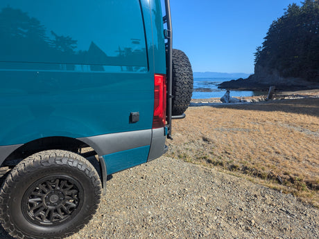 Teal RV rental van parked by a serene rocky beach with forest cliffs and blue ocean under a clear sky.