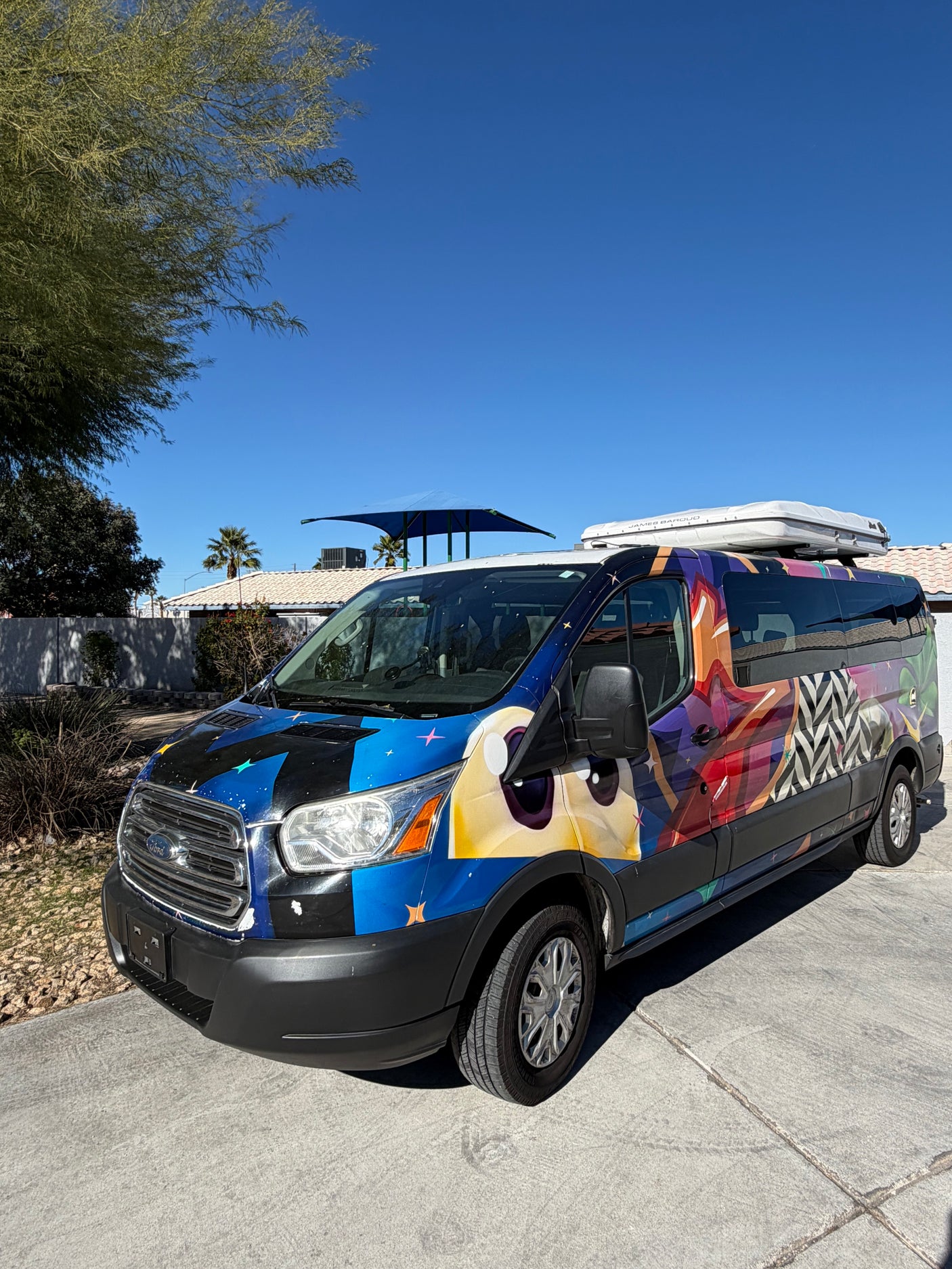 Vibrant graphic Ford Transit rv rental with a character art wrap & rooftop tent, parked on a sunny day.