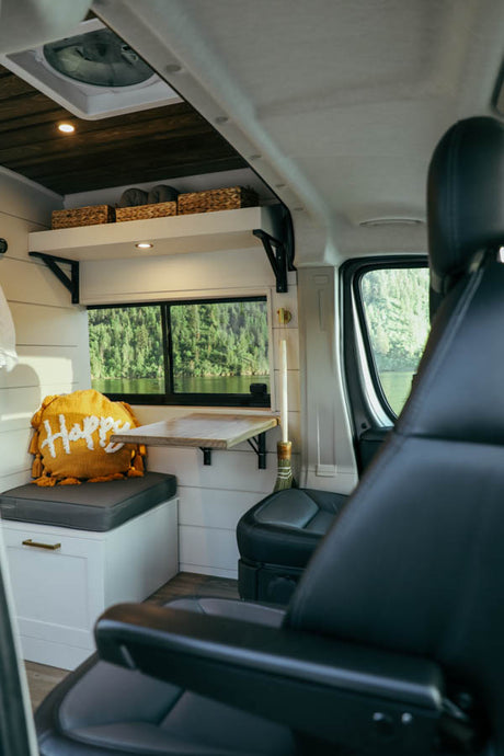 Cozy RV rental interior: white walls, wood ceiling, shelf, window with lake view, fold-down table, and comfy seating.