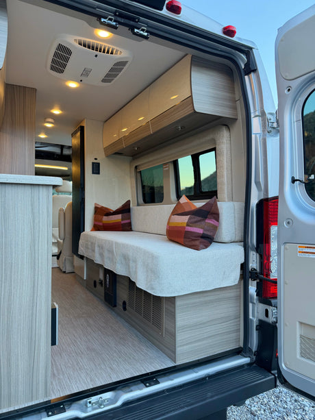 Inside a modern rv rental camper van, seen from open rear doors. Features a comfy bench, storage, AC, and bright interior.