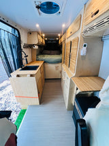 Bright RV rental van interior with light wooden cabinets, modern kitchen with black sink, rear bed, light floor, and organized storage.