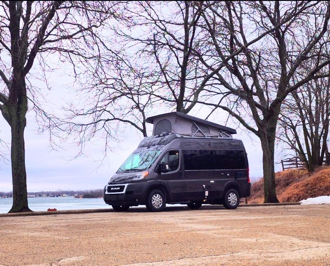 Dark gray Ram ProMaster campervan with pop-top for rv rental, parked lakeside among bare trees on an overcast day.