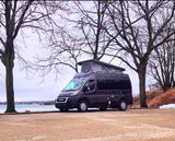 Dark gray Ram ProMaster campervan with pop-top for rv rental, parked lakeside among bare trees on an overcast day.