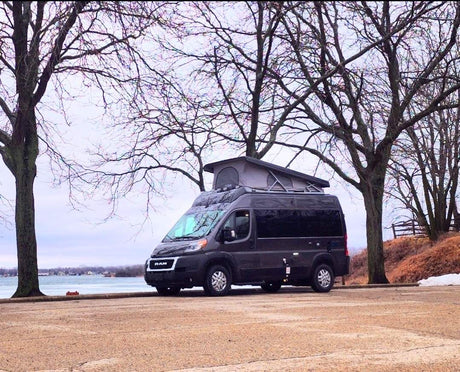 Dark gray Ram ProMaster campervan with pop-top for rv rental, parked lakeside among bare trees on an overcast day.