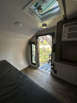 Inside an RV rental: compact living with a window view of trees, AC, and a cozy bed.
