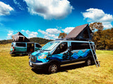 Two vibrant RV rental camper vans with rooftop tents open, parked in a sunny, grassy field amidst scenic mountains.