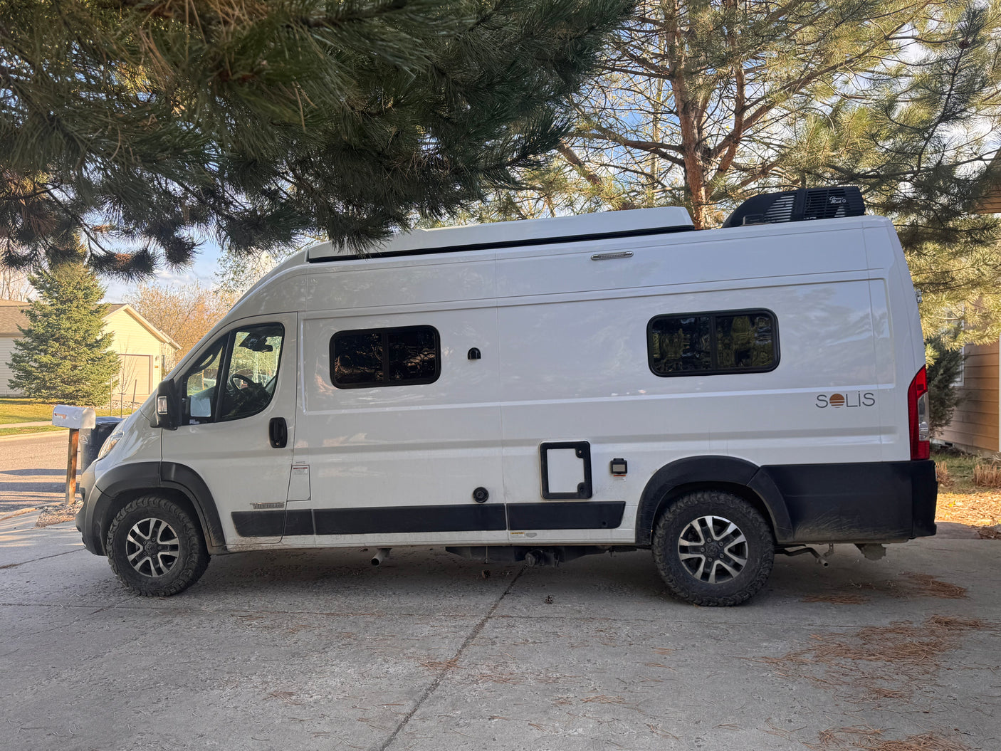 White Winnebago Solis camper van for rv rental, parked under a pine tree on a concrete driveway, ready for adventure.
