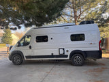 White Winnebago Solis camper van for rv rental, parked under a pine tree on a concrete driveway, ready for adventure.