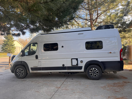 White Winnebago Solis camper van for rv rental, parked under a pine tree on a concrete driveway, ready for adventure.