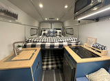 Spacious RV rental interior featuring a cozy bed with plaid bedding, compact kitchen with sink, induction cooktop, and microwave.