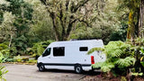 White camper van RV rental parked in a lush, green forest parking lot, surrounded by ferns and tall trees.