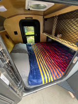 Cozy RV rental interior with a bed, colorful striped bedding, a window with outdoor view, and an overhead storage net.