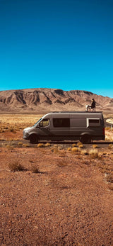 Road trip ready rv rental: A traveler relaxes on a van roof, surrounded by vast desert and rugged mountains under a clear blue sky.