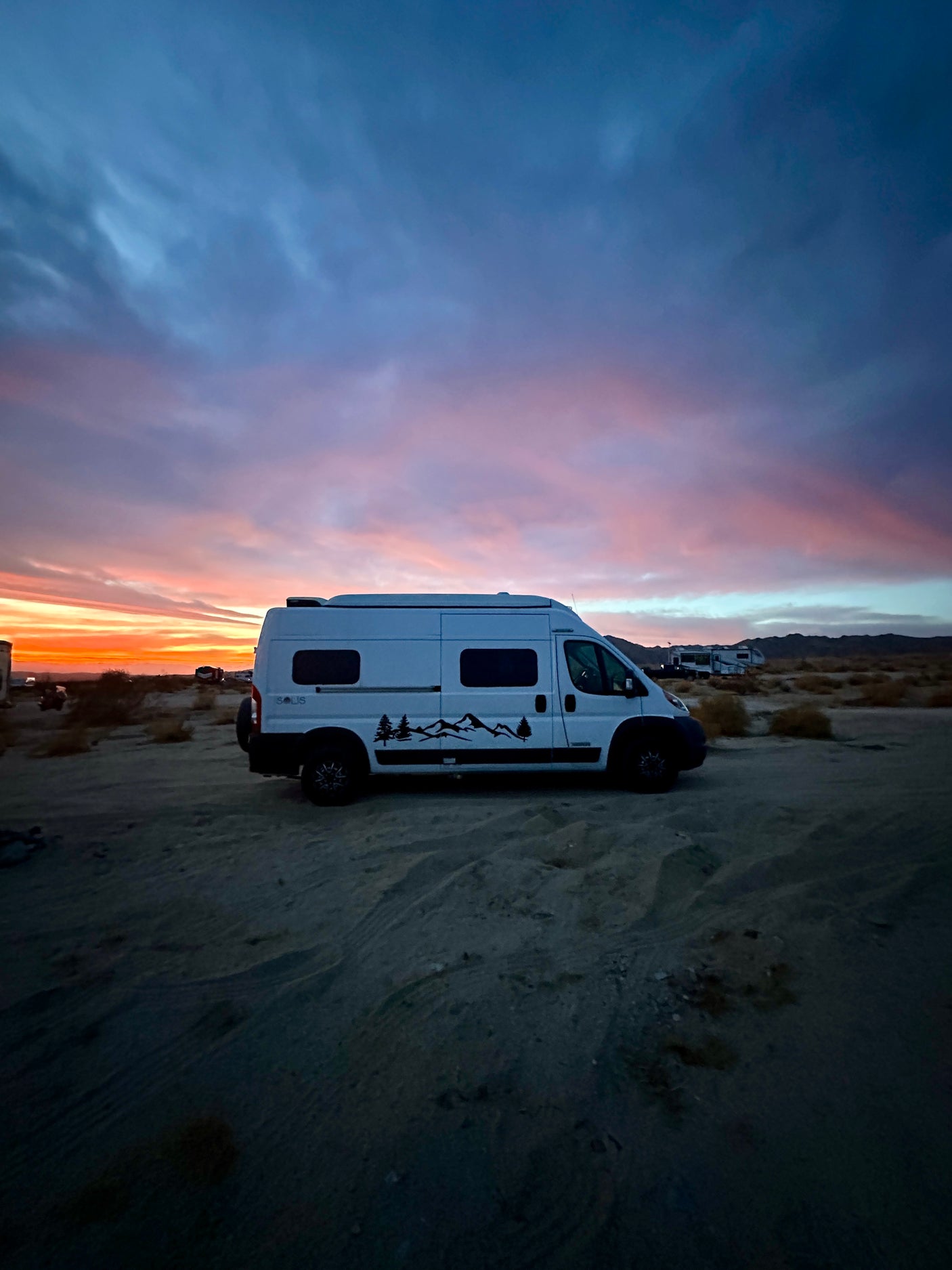 White rv rental camper van parked in a sandy desert under a vibrant sunset sky, perfect for outdoor travel.