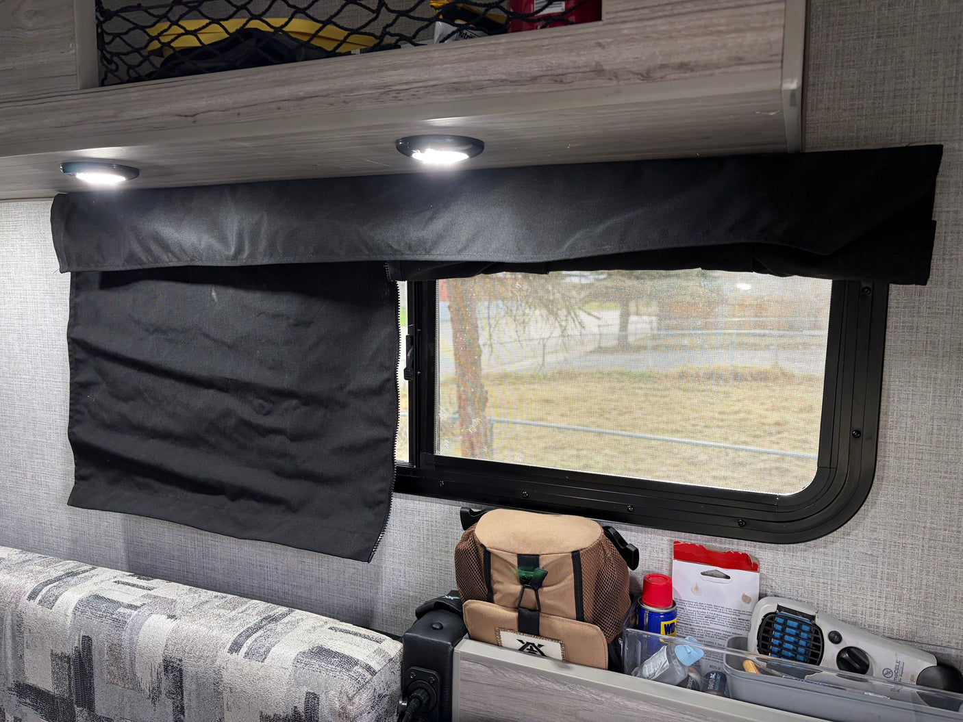RV rental interior shows a window with a black fabric shade, overhead lights, patterned sofa, and under-window storage for camping essentials like a Thermacell.
