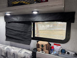RV rental interior shows a window with a black fabric shade, overhead lights, patterned sofa, and under-window storage for camping essentials like a Thermacell.