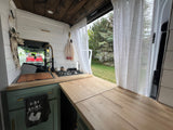 An RV rental's clever interior: compact kitchen with sink, stove, and extended counter space, framed by sheer curtains.