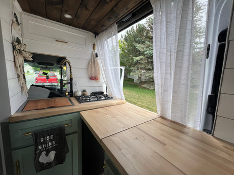An RV rental's clever interior: compact kitchen with sink, stove, and extended counter space, framed by sheer curtains.