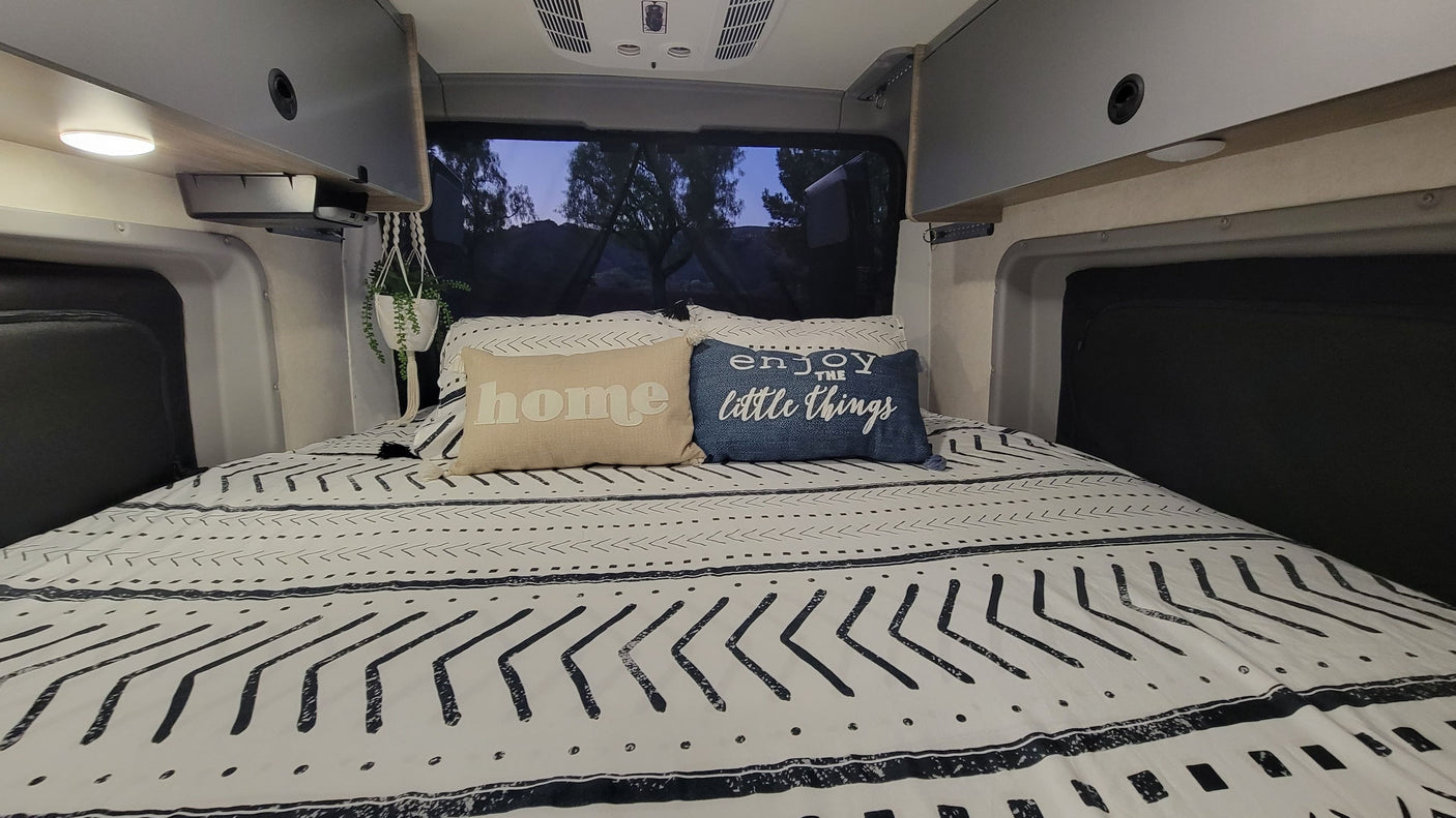 Cozy RV rental bed with black & white patterned bedding, 'home' & 'enjoy the little things' pillows, & overhead cabinets.