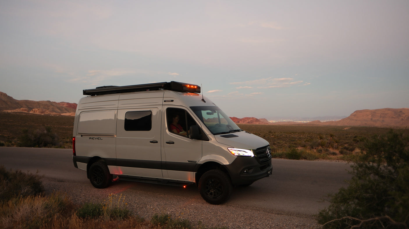 White Revel camper van on a desert road at dusk, lights glowing. Ideal for an rv rental adventure in scenic landscapes.