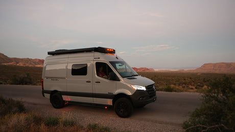 White Revel camper van on a desert road at dusk, lights glowing. Ideal for an rv rental adventure in scenic landscapes.