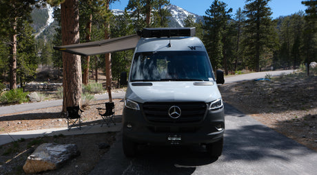 A grey Mercedes Sprinter RV rental with an extended awning and chairs is set up at a mountain campsite among pine trees.
