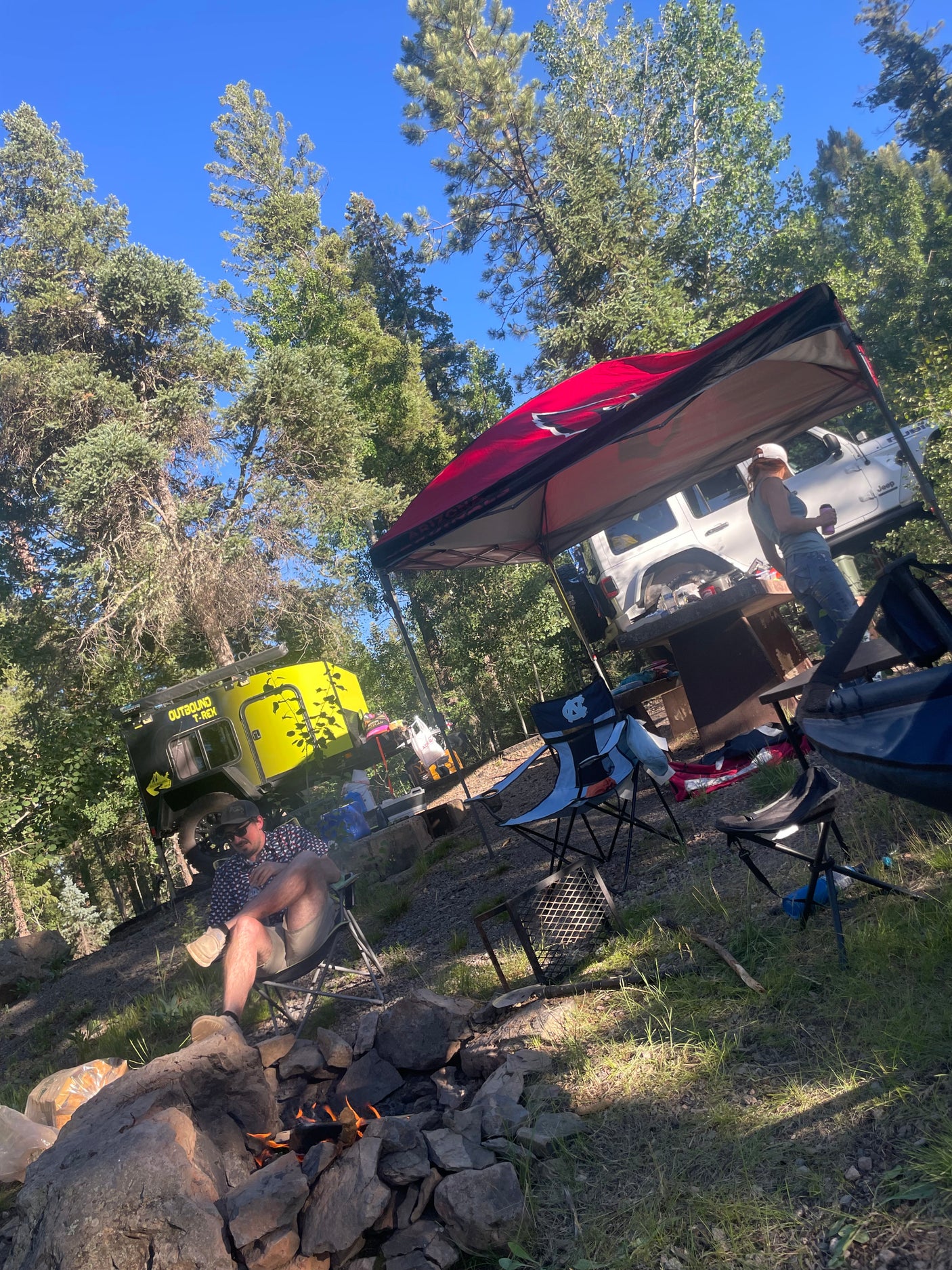 Forest camping scene with an Outbound T-Rex rv rental & Jeep. People relax by the campfire under a red canopy.
