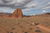 An RV rental drives on a red dirt road through a stunning desert landscape, passing massive red rock formations under a dynamic sky.