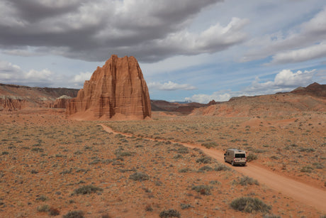 An RV rental drives on a red dirt road through a stunning desert landscape, passing massive red rock formations under a dynamic sky.