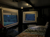 Cozy RV rental interior at dusk: two windows reveal a wooded view, complemented by a comfortable bed and soft ceiling lighting.