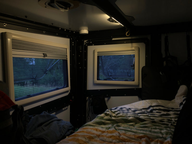 Cozy RV rental interior at dusk: two windows reveal a wooded view, complemented by a comfortable bed and soft ceiling lighting. — photo 3