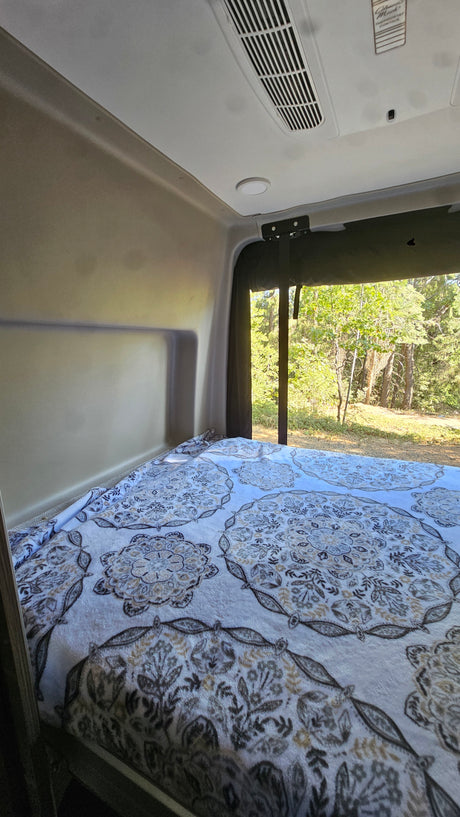 RV rental interior: cozy bed with a patterned blanket, looking out to a lush forest scene.