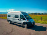 A silver Winnebago Solis RV rental van parked on a road, with mountains and green fields in the background.