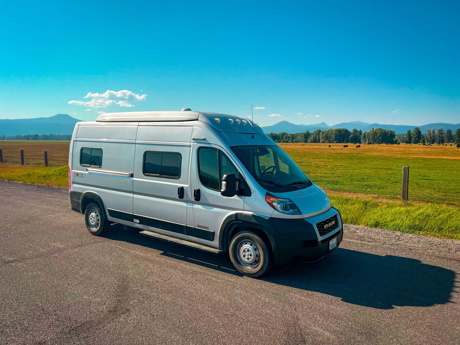A silver Winnebago Solis RV rental van parked on a road, with mountains and green fields in the background.