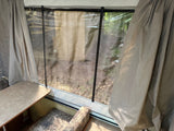 Inside an RV rental pop-up camper, looking out a window with clear plastic over mesh. Gray curtains, table, and seat are visible.