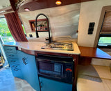 RV rental kitchen featuring light blue cabinets, a two-burner stove, microwave, sink, and wood accents with upholstered seating.