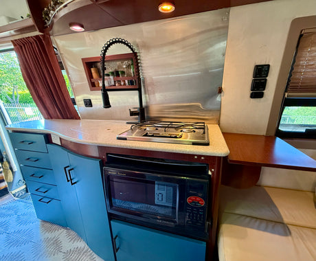 RV rental kitchen featuring light blue cabinets, a two-burner stove, microwave, sink, and wood accents with upholstered seating.