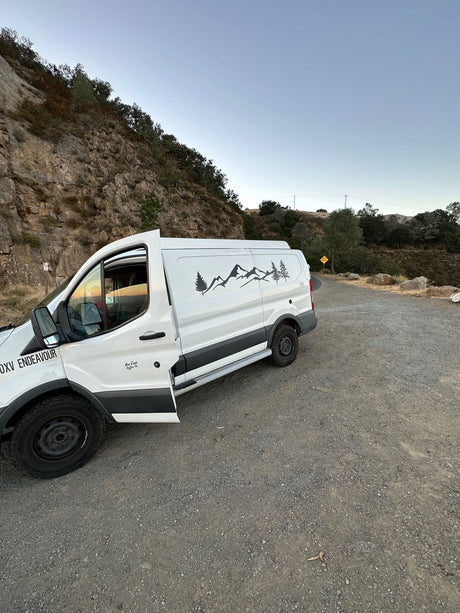 White camper van (RV rental) with mountain decals parked on a gravel road beside a rocky hillside.