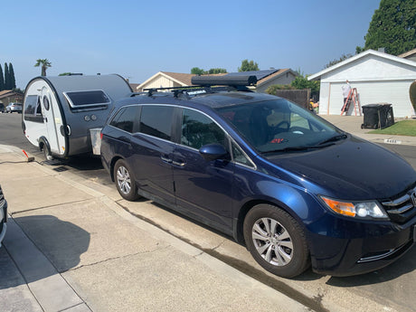 A blue minivan tows a compact grey travel trailer, ready for an RV rental adventure on a sunny street.