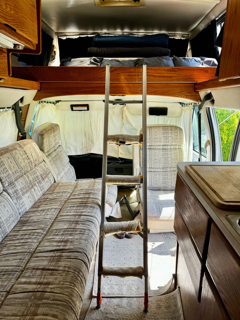  and living area with warm wood paneling.|Classic RV rental dashboard with a cassette player and a built-in shelf displaying a collection of audio tapes.|RV rental kitchen features a gas stove with three burners — photo 7